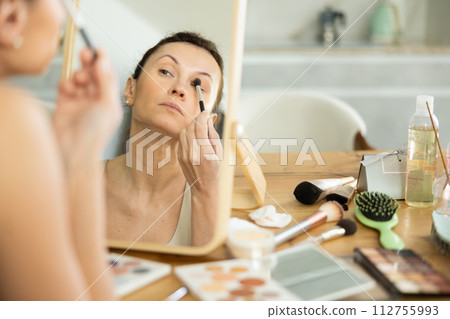 Middle-aged woman using brush during eye makeup sitting in front of the mirror Middle-aged woman using brush during eye makeup sitting in front of the mirror 112755993