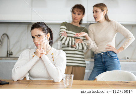 Upset middle-aged woman sitting at the kitchen table with her back to women quarreling to her 112756014