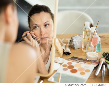 Woman reflected in tabletop mirror applying eyeliner during daily makeup Woman reflected in tabletop mirror applying eyeliner during daily makeup 112756085