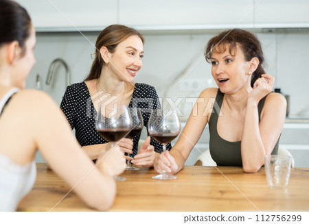 Three happy middle-aged women sitting at table with a glass of wine while having lively conversation Three happy middle-aged women sitting at table with a glass of wine while having lively conversation 112756299