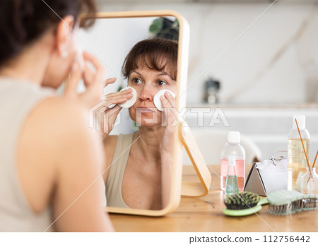 Middle-aged woman cleaning her facial skin with cotton disks sitting in front of the mirror 112756442