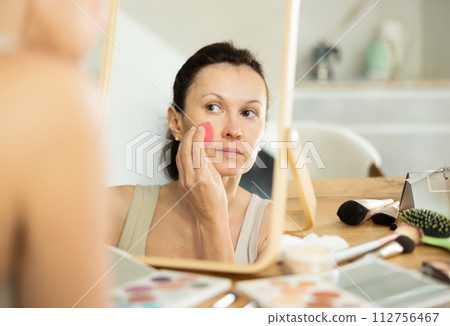 Middle-aged woman applying make-up foundation on her face by means of sponge 112756467