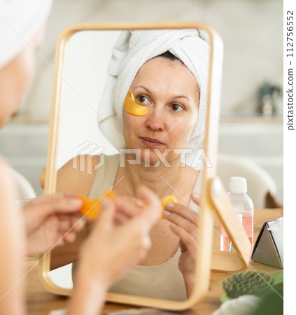Woman in towel turban applying golden collagen eye patches Woman in towel turban applying golden collagen eye patches 112756552