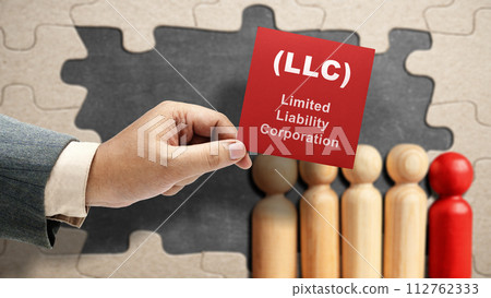 Business hand holding a note with LLC text, the acronym of Limited Liability Company with a row of wooden peg dolls 112762333