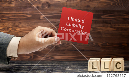 Business hand holding a note with LLC text, the acronym of Limited Liability Company with a row of wooden cubes with LLC text 112762337