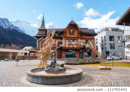 A serene Engelberg scene with a fountain sculpture, a Swissstyle building with a gabled roof, and contrasting modern architecture, set against snowcapped mountains and a clear sky. A serene Engelberg scene with a fountain sculpture, a Swissstyle building with a gabled roof, and contrasting modern architecture, set against snowcapped mountains and a clear sky. 112765443