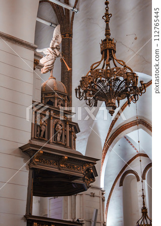 An ornate Riga Dome Cathedral interior with a gold accented wooden pulpit, white angel statue, and a large bronze chandelier, set against white walls and a red brick vaulted ceiling. An ornate Riga Dome Cathedral interior with a gold accented wooden pulpit, white angel statue, and a large bronze chandelier, set against white walls and a red brick vaulted ceiling. 112765445