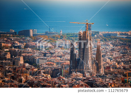 Panoramic view of Barcelona featuring the unfinished Sagrada Familia with its spires and cranes, surrounded by the city's warmhued buildings and the Mediterranean Sea in the backdrop. 112765679