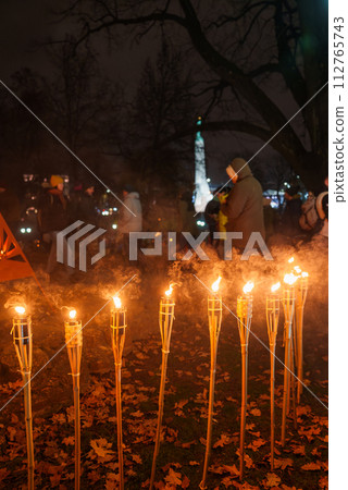 Nighttime celebration in Latvia with torches lighting up fallen leaves and a crowd in winter attire, creating a warm, traditional atmosphere in an outdoor setting. 112765743