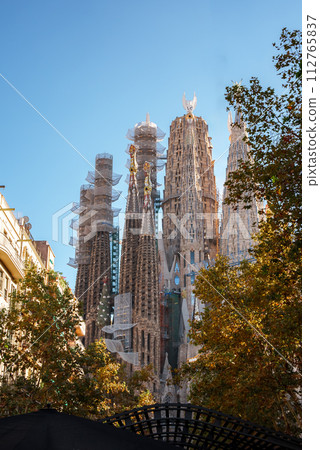 Autumn at Barcelona's Sagrada Familia reveals its Gothic and Art Nouveau splendor against a clear blue sky, framed by colorful leaves, with ongoing construction evident by cranes. Autumn at Barcelona's Sagrada Familia reveals its Gothic and Art Nouveau splendor against a clear blue sky, framed by colorful leaves, with ongoing construction evident by cranes. 112765837