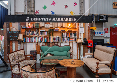 A rustic, homely indoor seating area with a vintage green sofa, floral armchair, and wooden chairs at Vaerftets Madmarked, featuring a bookshelf and colorful decor. 112765860