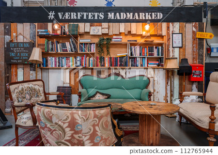 A cozy, eclectic indoor seating area with a green sofa, floral armchair, and rustic table, surrounded by bookshelves and wooden decor, likely in a Helsingborg food market. 112765944