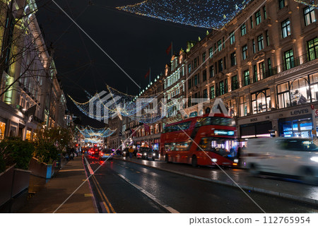 A bustling London Regent street at Christmas time, adorned with festive lights that mimic a starry sky. The scene includes iconic red buses, lively shops, and a warm, celebratory atmosphere. A bustling London Regent street at Christmas time, adorned with festive lights that mimic a starry sky. The scene includes iconic red buses, lively shops, and a warm, celebratory atmosphere. 112765954