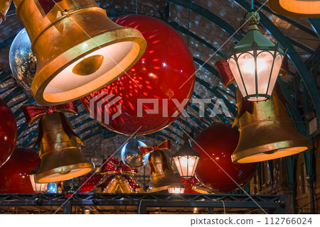 Covent Garden indoor decorations featuring oversized red baubles and golden bells hang from the arched ceiling of a traditional London market, creating a warm, celebratory Christmas atmosphere. Covent Garden indoor decorations featuring oversized red baubles and golden bells hang from the arched ceiling of a traditional London market, creating a warm, celebratory Christmas atmosphere. 112766024