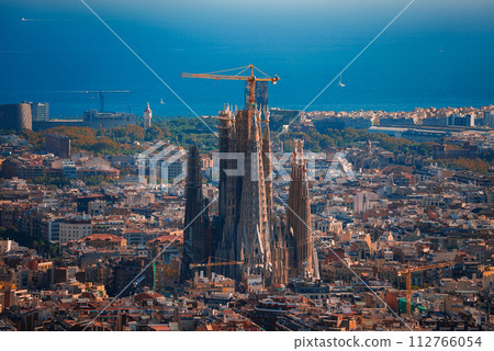 Panoramic view of Barcelona featuring the unfinished Sagrada Familia with its towering spires and cranes. The city's Mediterranean architecture and the blue sea horizon under a clear sky are visible. 112766054