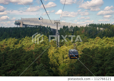 A serene cable car ride at Druskininkai resort in Lithuania, with DRUSKININKAI branding, offers panoramic views of a green forest and a modern terminal building. 112766121