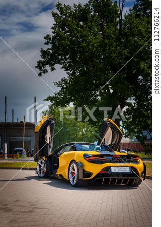 A bright yellow McLaren 765LT, with dihedral doors open, is displayed outside. Black details accent its sporty look. The back has circular taillights and quad exhaust. 112766214