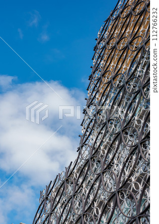 A closeup of a modern building in Birmingham, UK, with a facade featuring interlinked metallic circles under a partly cloudy blue sky, reflecting silvery to golden tones. 112766232