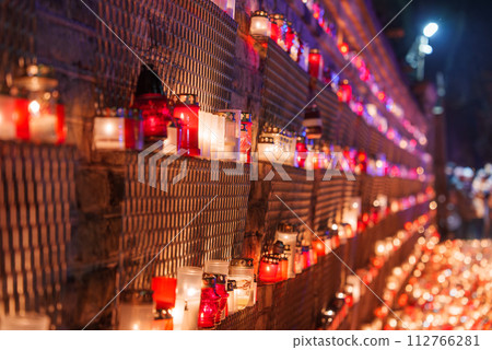 Closeup of red and white candles lit in rows on a mesh wall, creating a reflective ambiance for Latvia's Independence Day, likely in Riga or a significant monument. 112766281