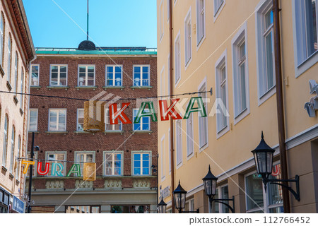 Europeanstyle buildings line a serene street under a clear sky, with colorful letters spelling KAKA and TURA strung overhead, and classic lanterns adding charm, possibly in Helsingborg. 112766452