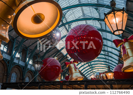 Inside a historic London venue, Covent Garden Market, oversized red baubles with golden bows and bellshaped lights create a warm, festive atmosphere under a curved glass roof. Inside a historic London venue, Covent Garden Market, oversized red baubles with golden bows and bellshaped lights create a warm, festive atmosphere under a curved glass roof. 112766490