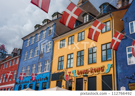 Vibrant Copenhagen street scene with colorful European buildings in blue, yellow, and red, Danish flags waving, and signs for a Hong Kong shop and a cafe. Vibrant Copenhagen street scene with colorful European buildings in blue, yellow, and red, Danish flags waving, and signs for a Hong Kong shop and a cafe. 112766492