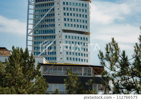 A modern skyscraper, the Turning Torso in Malmo, Sweden, twists skyward with a white facade and window pattern, framed by evergreens on a clear day with blue skies. A modern skyscraper, the Turning Torso in Malmo, Sweden, twists skyward with a white facade and window pattern, framed by evergreens on a clear day with blue skies. 112766575