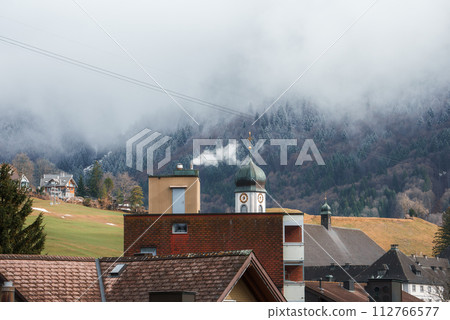 Scenic view of a mountain village in Engelberg with a clock tower, smoke from a chimney, and misty forested slopes, suggesting a serene, chilly ambiance before ski season. 112766577