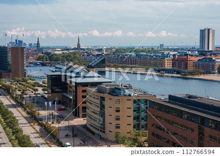 Daytime cityscape from an elevated view, featuring modern midrise buildings with brick and glass facades, a treelined street, and a reflective water body with boats in Copenhagen, Denmark. 112766594