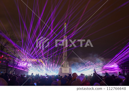 A lively Independence Day celebration in Latvia with a laser light show casting purple and pink beams across the night sky, centered around Riga's iconic Freedom Monument. A lively Independence Day celebration in Latvia with a laser light show casting purple and pink beams across the night sky, centered around Riga's iconic Freedom Monument. 112766648