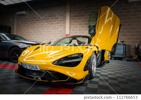 A vibrant yellow McLaren 765LT with upwardopening dihedral doors is displayed in a tuning garage, featuring black aerodynamic accents and multispoke wheels against a checkered floor. 112766653