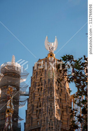 Closeup of Sagrada Familia's tower in Barcelona, adorned with Gaudi's signature motifs, a backlit cross, and dove sculptures against a clear sky, framed by foliage. 112766698