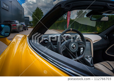 View from the driver's side into a yellow McLaren 765LT's luxurious interior, featuring a leatherwrapped steering wheel with beige stitching, and a hightech console. 112766703
