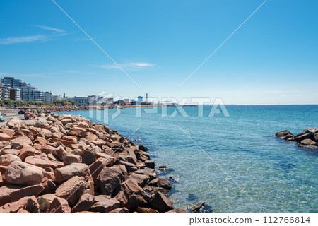 A serene coastal landscape with a rocky shoreline and calm waters under a clear blue sky. Modern buildings line the coast, suggesting an urban beachfront in Helsingborg. 112766814