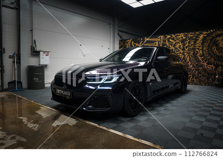 A sleek black BMW with headlights on is centered in a tuning garage with a checkered floor, wet patches reflecting light, and walls featuring edgy graffiti, indicating a highperformance service area. 112766824