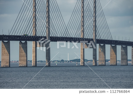 A modern suspension bridge with tall pylons and fanning cables spans calm waters under an overcast sky, likely the Oresund Bridge connecting Copenhagen and Malmo. A modern suspension bridge with tall pylons and fanning cables spans calm waters under an overcast sky, likely the Oresund Bridge connecting Copenhagen and Malmo. 112766864