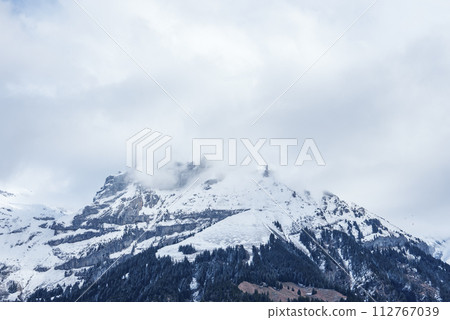 Majestic snowcovered mountains rise in Engelberg, Switzerland, with rugged peaks shrouded by clouds. Foreground hints at wooded foothills under an overcast sky, capturing the serene alpine beauty. 112767039