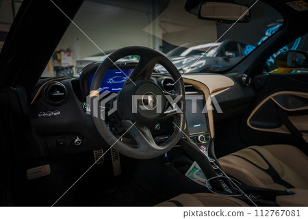 Inside a McLaren 765LT, the image showcases a sporty steering wheel with the McLaren logo, control buttons, and a digital dashboard display. A twotone interior and hightech center console add luxury. Inside a McLaren 765LT, the image showcases a sporty steering wheel with the McLaren logo, control buttons, and a digital dashboard display. A twotone interior and hightech center console add luxury. 112767081