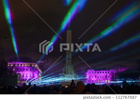 Nighttime celebration in Riga, Latvia, with silhouetted onlookers admiring the illuminated Freedom Monument amid a vibrant light show of green, blue, and pink beams, flanked by classical buildings. 112767087
