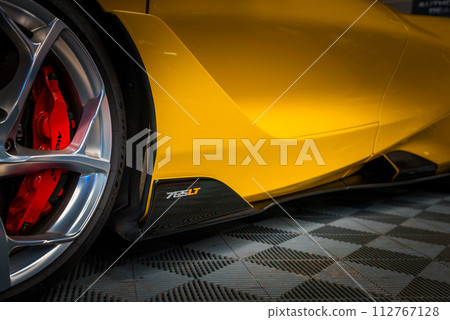 Closeup of a yellow McLaren 765LT's sleek side profile near the rear wheel, highlighting its aerodynamic design, red brake caliper, and silver alloy wheel on a checkered floor. Closeup of a yellow McLaren 765LT's sleek side profile near the rear wheel, highlighting its aerodynamic design, red brake caliper, and silver alloy wheel on a checkered floor. 112767128