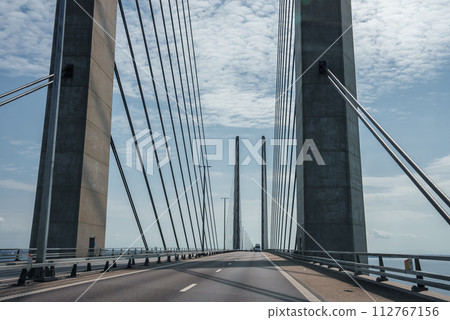 Capturing the sleek engineering of the Oresund Bridge, this image features its towering concrete supports and harplike cables under a clear blue sky, on a trafficfree multilane highway. 112767156