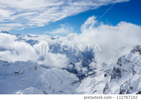 Breathtaking winter landscape of Engelberg, Switzerland, with snowcovered mountains under a bright blue sky dotted with fluffy clouds, perfect for skiing and snowboarding. 112767182