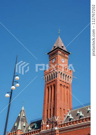 A red brick Gothic Revival tower with a clock, spire, and arched windows stands under a clear sky, beside a street lamp and a historic building, possibly in Helsingborg. 112767202