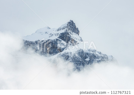 A snowcovered mountain peak rises above clouds in Engelberg, Switzerland, with a serene mix of whites and grays, highlighting the rugged, challenging terrain. A snowcovered mountain peak rises above clouds in Engelberg, Switzerland, with a serene mix of whites and grays, highlighting the rugged, challenging terrain. 112767321