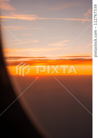 Capturing a serene sunset from an airplane window, the sky displays a gradient of warm colors with a fiery sun partially hidden by the horizon and scattered clouds highlighted by its glow. 112767339