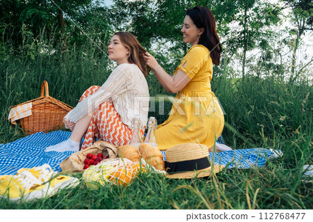 A young woman braids her friend's hair at a picnic in the park. A young woman braids her friend's hair at a picnic in the park. 112768477