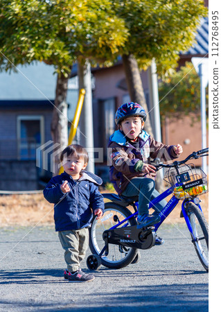 A boy riding a bicycle in the park and his younger brother following behind him A boy riding a bicycle in the park and his younger brother following behind him 112768495
