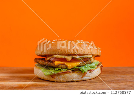 Delicious cheeseburger with lettuce, tomato, cheese, and bacon on a wooden surface. Studio shot with orange background. Fast food and indulgence concept. Design for menu, poster, and banner Delicious cheeseburger with lettuce, tomato, cheese, and bacon on a wooden surface. Studio shot with orange background. Fast food and indulgence concept. Design for menu, poster, and banner 112768577
