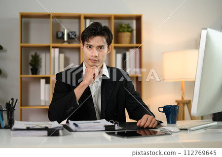 Confident millennial businessman sitting at workplace and looking at camera Confident millennial businessman sitting at workplace and looking at camera 112769645