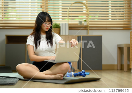 Young woman sitting on on exercise mat and using laptop computer for watching online tutorial Young woman sitting on on exercise mat and using laptop computer for watching online tutorial 112769655
